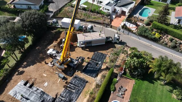 Pre-fabricated modular house during unloading operations from truck in construction area. Aerial top-down orbiting