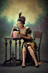 Portrait of young beautiful girl, princess in elegant dress wearing 3D glasses, eating popcorn and watching movie over dark vintage background. Concept of history, renaissance art, comparison of eras