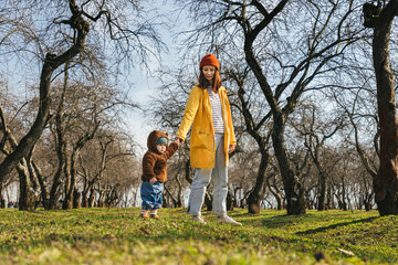 a young mother holds a small child by the hand who takes the first steps in the park, an alley of trees without leaves