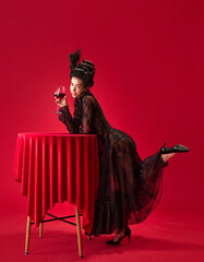 Portrait of beautiful, attractive young girl in elegant, stylish, black dress leaning on table, holding glass with red wine against red background. History, renaissance art, comparison of eras concept