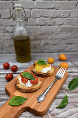 Tasty bruschetta with ricotta cheese, red and yellow cherry tomatoes and basil on a wooden cutting board. Ingredients on the background, vertical image with bokeh effect.