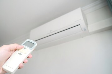 Modern air conditioner on white wall indoors. Hand with remote control directed on air conditioner.