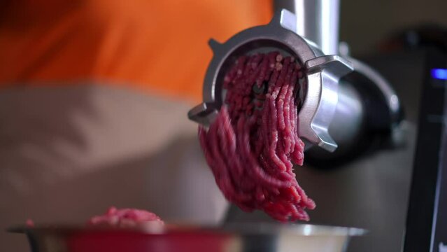 Close-up meat grinder with ground meat falling out of equipment in restaurant kitchen. Unrecognizable female Caucasian cook preparing raw ingredient for cooking indoors