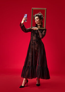 Full-length portrait of tender, beautiful, young girl holding picture frame and taking selfie with phone against red background. Concept of history, renaissance art, comparison of eras