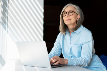 Pensive 60 years old businesswoman, successful gray haired manager using laptop computer, planning...