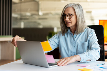 Attractive gray haired senior business woman wearing eyeglasses using laptop computer, working online sitting in modern office, Successful manager using sticky notes, agile methodology. Scrum concept