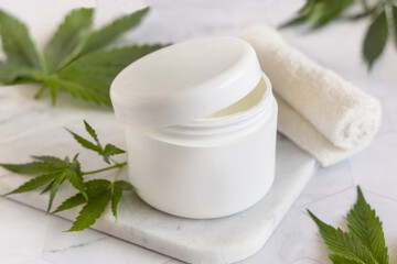 Fototapeta na wymiar Opened white cream jar with a lid near cannabis leaves. Cosmetic Mockup