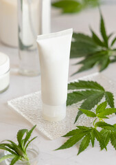 Obraz na płótnie Canvas Blank Cream tube near green cannabis leaves on white table. Cosmetic Mockup
