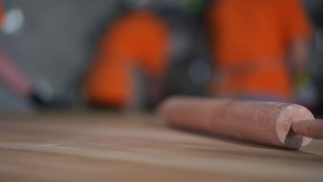 Close-up rolling pin on wooden tabletop on the right with blurred cooks working in restaurant at background. Unrecognizable Caucasian women cooking in cafe indoors