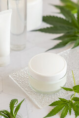 White cream jar with a blank lid near cannabis sativa leaves. Cosmetic Mockup