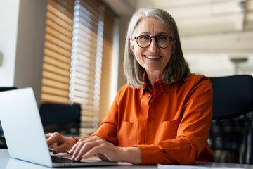 Portrait of smiling confident senior businesswoman wearing stylish eyeglasses, using laptop...