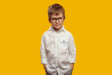 Fototapeta premium Serious little boy wearing white shirt and eyeglasses, keeping hands in pockets and looking mad at camera against yellow background