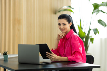 Young woman holding a tablet pc while sitting behind her desk in the office with a smile