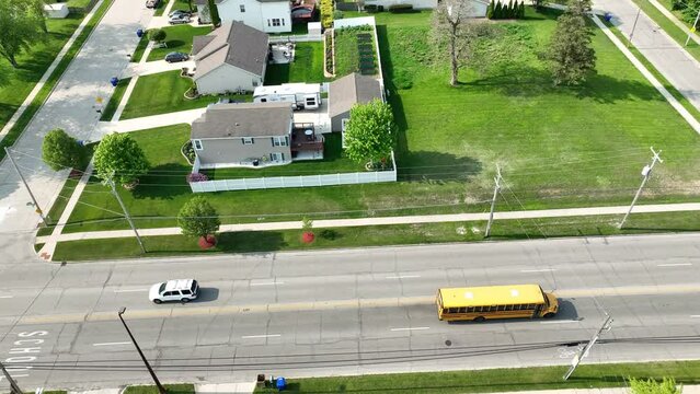 School bus driving down a residential street with school painted on the road from an Aerial view. The road is lined with houses, open green grass areas and trees.
