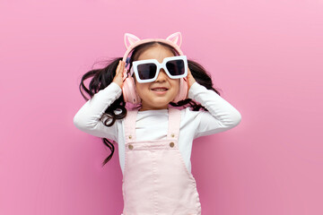 little asian girl in pink children's headphones and glasses listens to music and dances on pink...