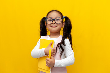 little asian girl with glasses holding books and smiling over yellow isolated background, korean smart preschool child - obrazy, fototapety, plakaty