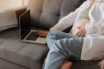 Young pregnant woman caress her big belly and remotely works on laptop at home on sofa, communicating online, browsing internet social media. Business, pregnancy and Internet technology concept.