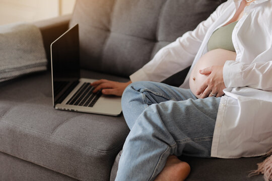 Young pregnant woman caress her big belly and remotely works on laptop at home on sofa, communicating online, browsing internet social media. Business, pregnancy and Internet technology concept.