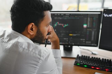 Side view of smart Indian crypto trader, investor, analyst broker, using laptop and smartphone analyzing digital cryptocurrency exchange, stock market charts, thinking of investing and funds risks.