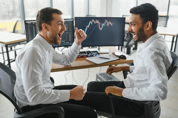 Two men traders sitting at desk at office together monitoring stocks data candle charts on screen...