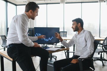Two men traders sitting at desk at office together monitoring stocks data candle charts on screen analyzing price flow smiling cheerful having profit teamwork concept