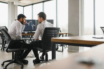 Two men traders sitting at desk at office together monitoring stocks data candle charts on screen analyzing price flow smiling cheerful having profit teamwork concept