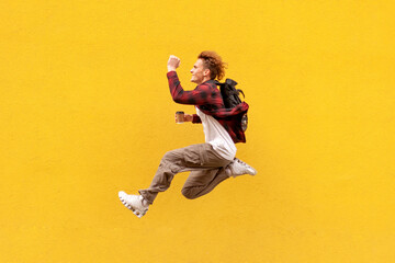 young cheerful guy student with backpack and coffee quickly runs in the air and jumps against the...