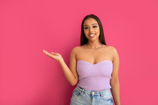 Cheerful young lady in fashionable top pointing at copy space on pink studio background. Smiling woman showing some advertisement