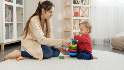 Cute baby boy assembling colorful toy tower with his mother on floor in living room