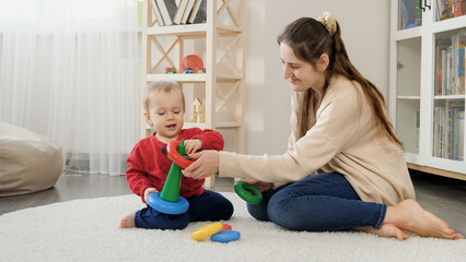 Little baby with mother learning assembling colorful toy pyramid on floor in living room. Baby development, child playing games, education and learning.