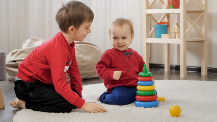 Smiling older boy playing with his little baby brother in toys on carpet at living room