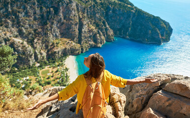 Traveler girl on vacation sitting on cliff, freedom opening arms, enjoying amazing panoramic view of Butterfly Valley Beach - 605715439