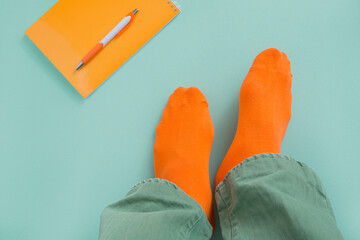 Feet of a man in orange socks and green jeans. Teal background with notepad and pen