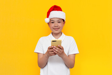 little asian boy in santa hat uses smartphone on yellow isolated background, korean child in new year's hat