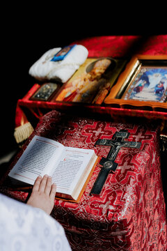 Hand of priest holds opened Holy Bible on the lectern near cross and icons.