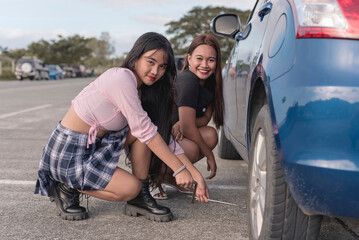 Two pretty ladies on the side of the road trying to help each other in using the jack to lift the sedan car.