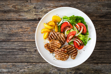 Grilled pork loin steaks with fresh vegetable salad and fried potatoes on wooden table
