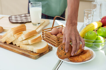 Close-up hand young asian man reaching for cookie placed in plate on kitchen table, grabbed a cookie ate it pick up glass milk before walking out kitchen, In other hand, holds laptop computer work.