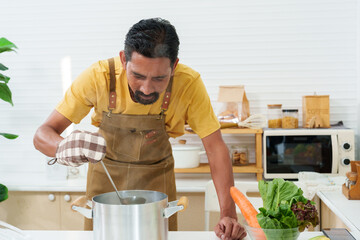 Asian man wearing an apron prevent while cooking in kitchen, wearing heat-resistant gloves as was cooking soup in large soup pot, keep holding ladle stir soup lowered head near pot inhale aroma soup.
