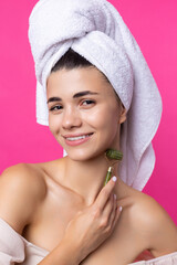 A beautiful cheerful attractive girl with a towel on her head holds a cosmetic roller near her face against a pink background.