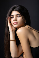 Fototapeta premium Classic studio portrait of a young brunette dressed in a black top, who is sitting on a chair against a black background.
