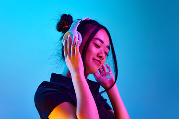 Portrait of beautiful young korean girl listening to music in headphones against blue studio...