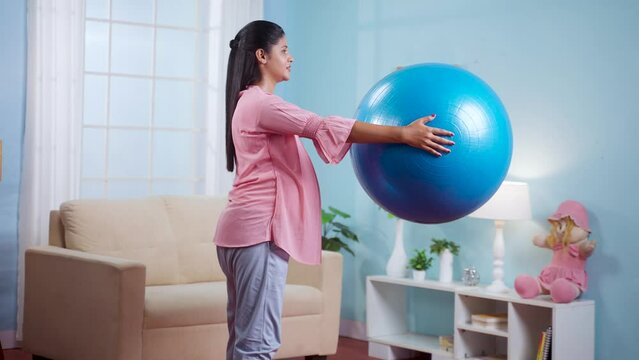 Indian pregnant woman doing squats exercise by hoding fitness ball at home - conept of Maternity workout, active lifestyle and Endurance.