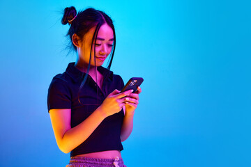 Portrait of young korean girl smiling and typing message on phone against blue studio background in...