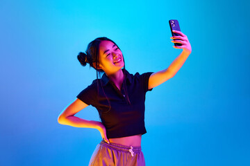 Naklejka premium Portrait of smiling, young, korean girl taking selfie with mobile phone against blue studio background in neon light. Concept of emotions, facial expression, youth, lifestyle, inspiration, sales, ad