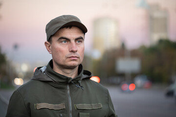 Portrait of serious military man in olive uniform and cap walking in city street on sunset