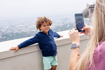 Cute diverse and confident little boy smiling while his mother takes his picture at a picturesque and scenic viewpoint about a large cityscape. Smart phone photography concept photo