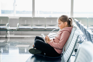 Kid, teen girl waiting for plane flight in departure hall. Sitting on chairs, playing computer games in gadgets, mobile phone, tablet. Chatting in social networks, wathing video. Study online