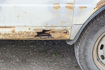 part of an old white metal car with brown rust and a hole at the black wheel in the street