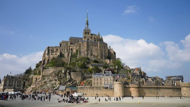 Tourists Walking Around By Le Mont Saint-Michel Castle On Sand Of Low Tide On Sunny Day On Background Of Blue Sky. Crowd Of People Strolling On Sand Sea, Seagulls Flying Though Air, Slow Motion.